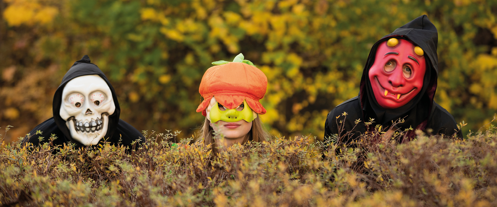 3 personer i halloweenmasker tittar upp bakom en buske.