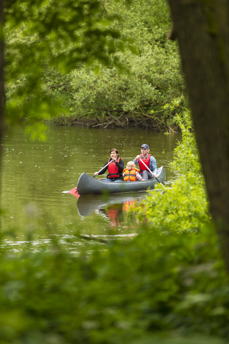 Två vuxna och ett barn sitter i en kanot. De verkar paddla på en å, och omgivningarna är lummiga och gröna.