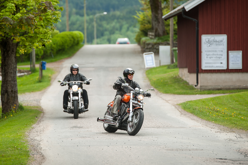 En bild på två män på varsin motorcykel som åker på en asfalterad väg omgiven av naturens grönska