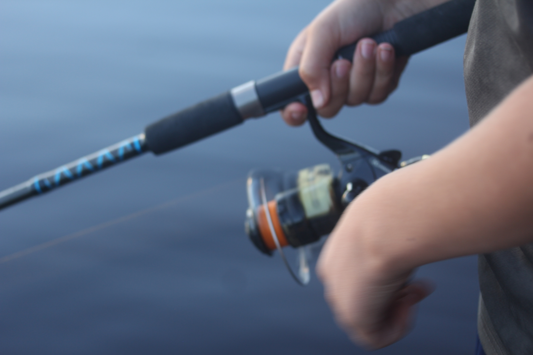 En närbild på två händer som håller i ett fiskespö. I bakgrunden kan man se vattenytan på en sjö.