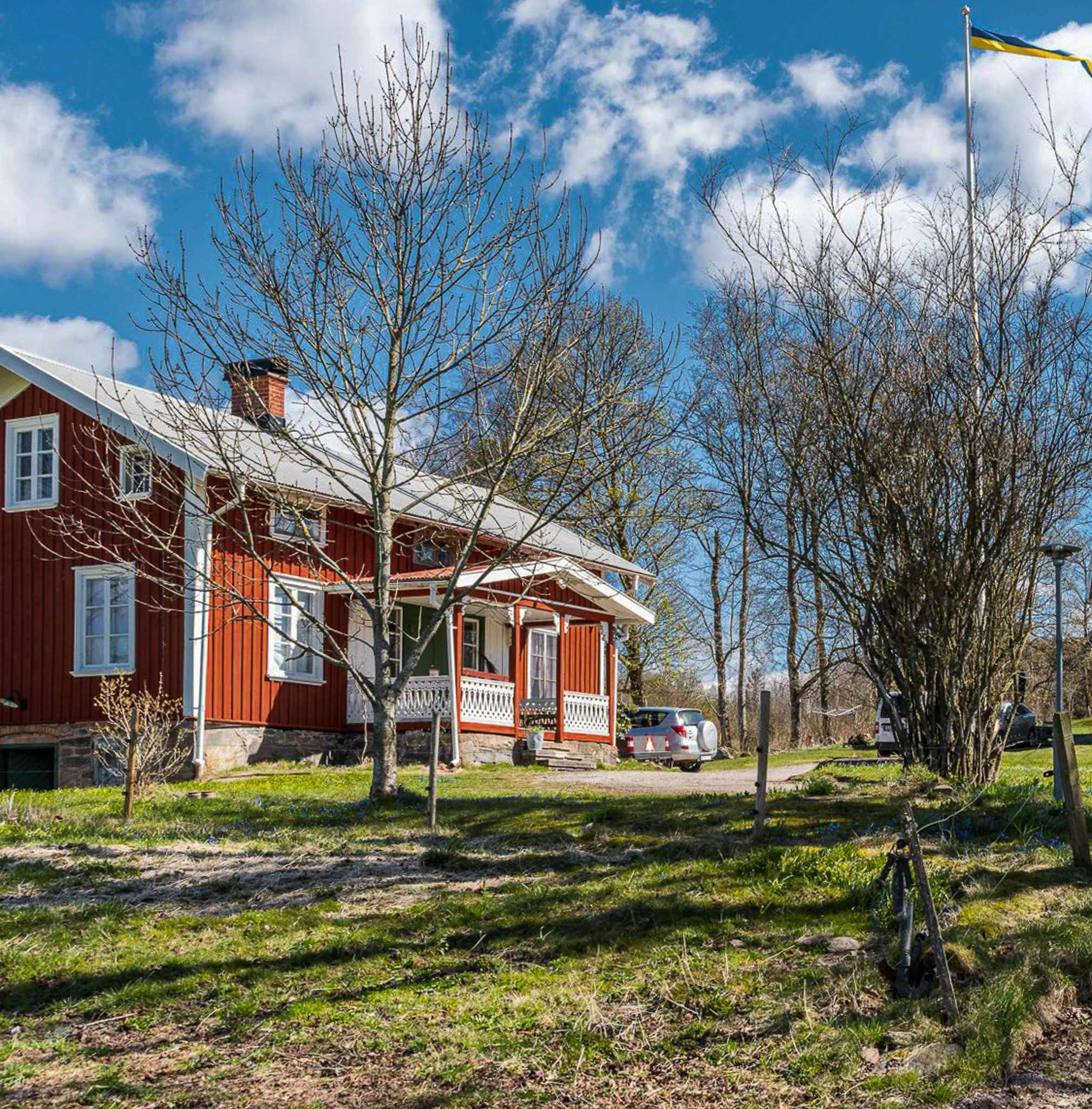 En bild på ett äldre rött hus med en stor vitmålad veranda. Framför huset är en gräsmatta med några träd.