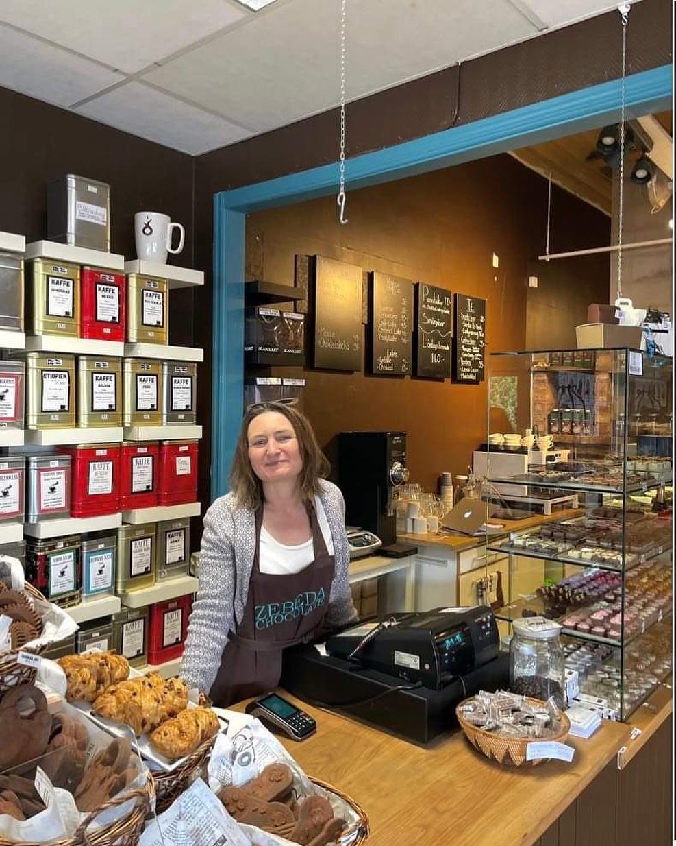 En bild på en kvinna iklädd förkläde som står bakom en cafédisk inne mysigt café som går i bruna toner.