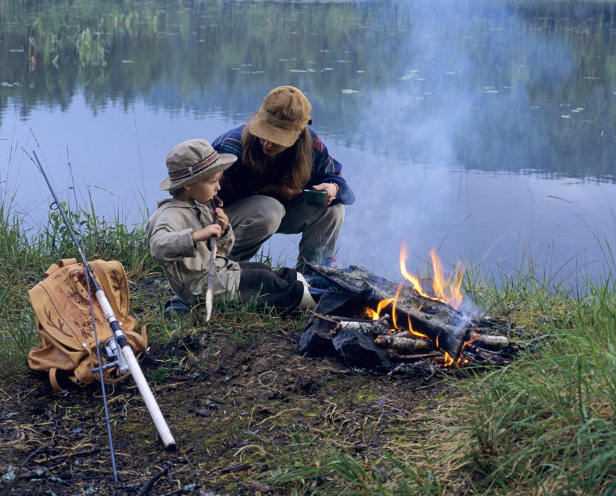 En bild på en kvinna och ett barn som sitter vid en lägereld jämte en sjö och grillar korv. Jämte dem ligger en ryggsäck och ett fiskespö.