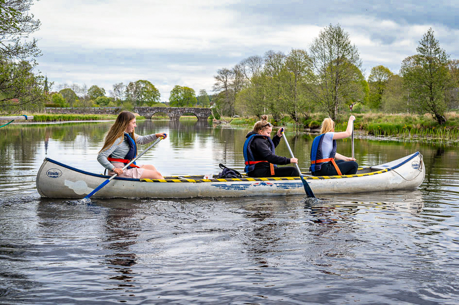 Tre ungdomar paddlar kanot i en å. I bakgrunden ser man en gammal stenbro. Längs kanterna på ån är det grönt och frodigt.