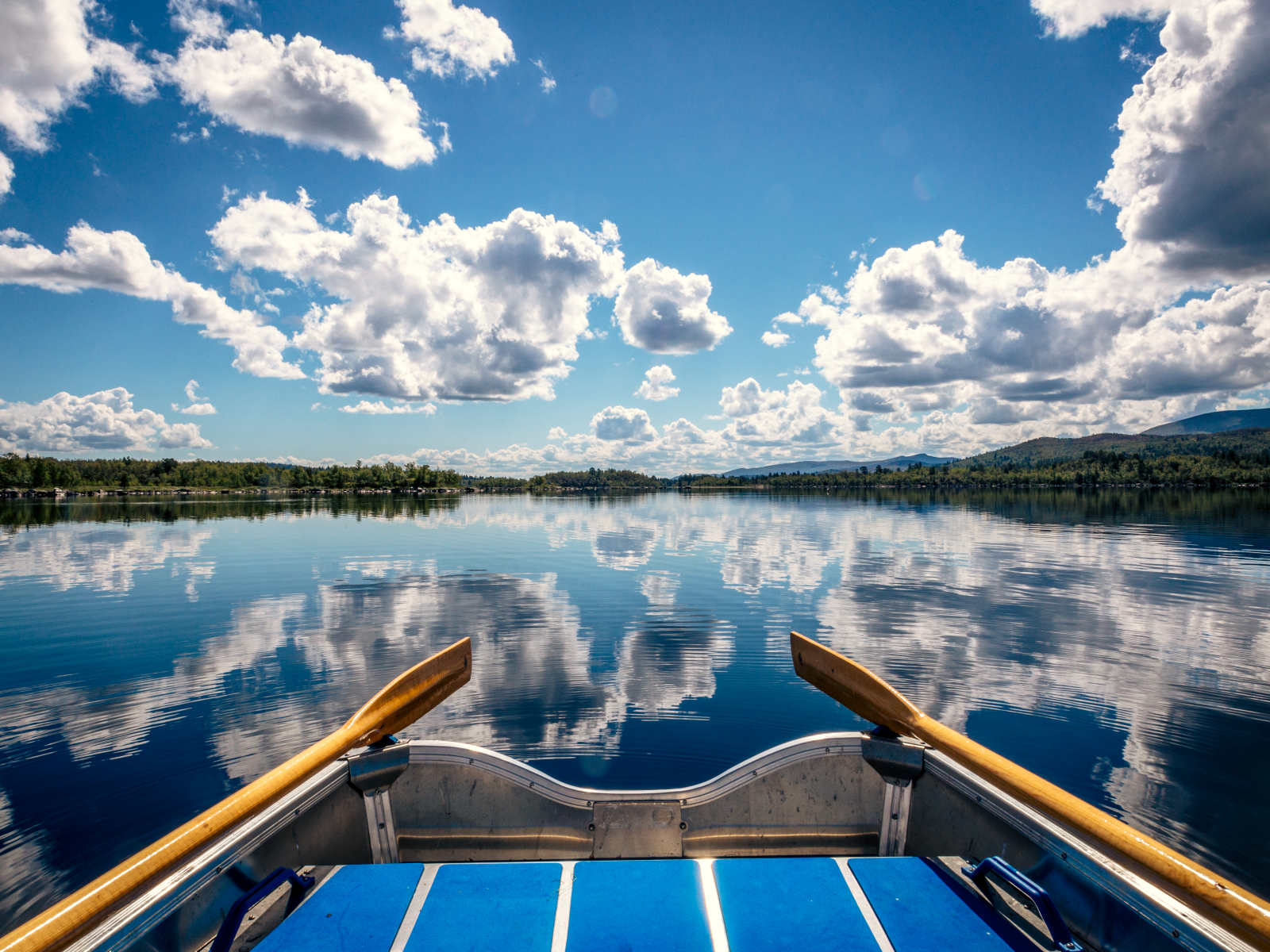 Bilden är tagen från en roddbåt och man skymtar en liten del av båten samt årorna, men mest fokus är det på den stilla sjön där himlen speglar sig.