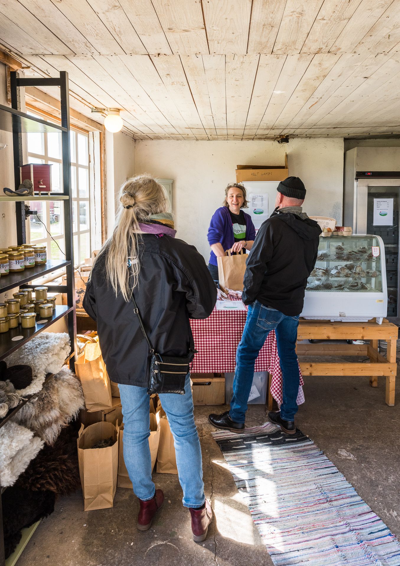 En bild på två människor som står och handlar av en kassörska i en liten gårdsbutik.