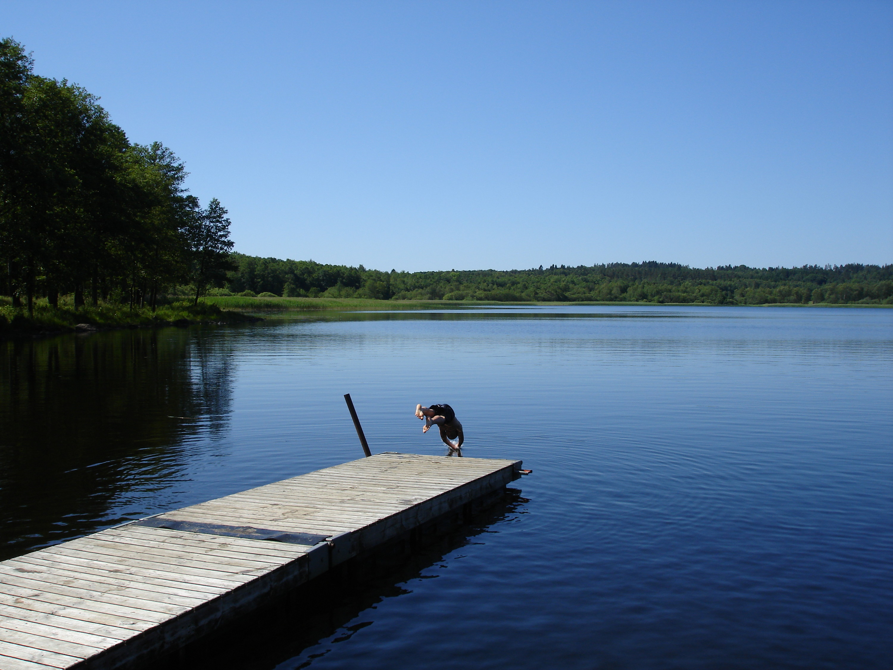En bild på en blå sjö med en brygga där någon dyker ner i vattnet.