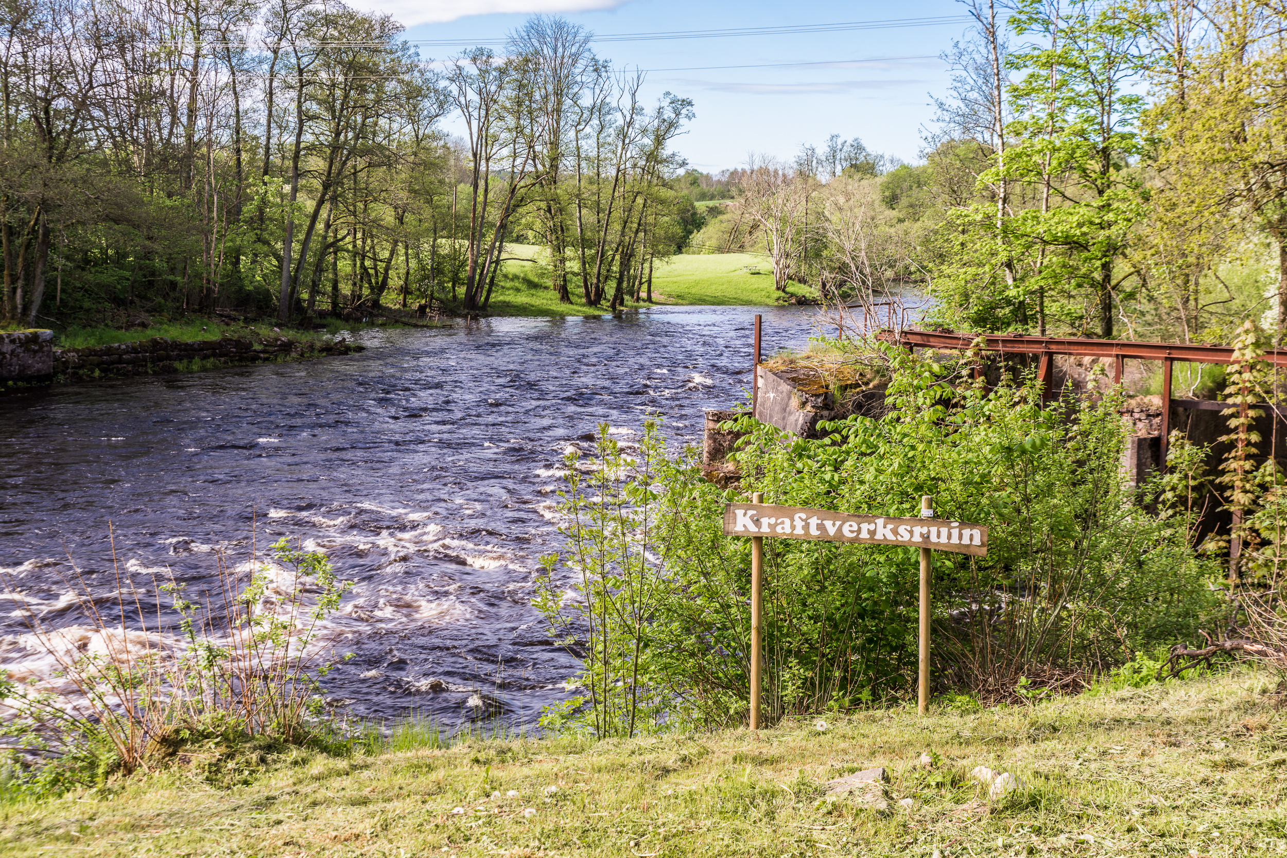 En bild på en strömmande å i ett grönt landskap. Vid ån sitter en träskylt med texten "Kraftverksruin".