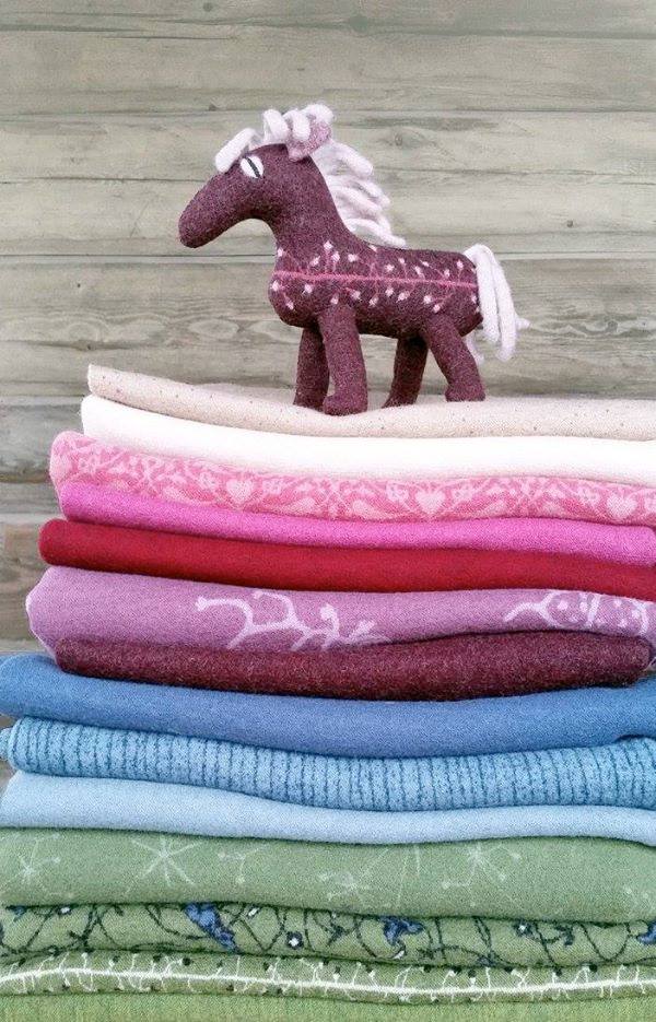 En bild på vikta tyger av ull som lagts i en hög. På toppen står en häst av tovad ull.