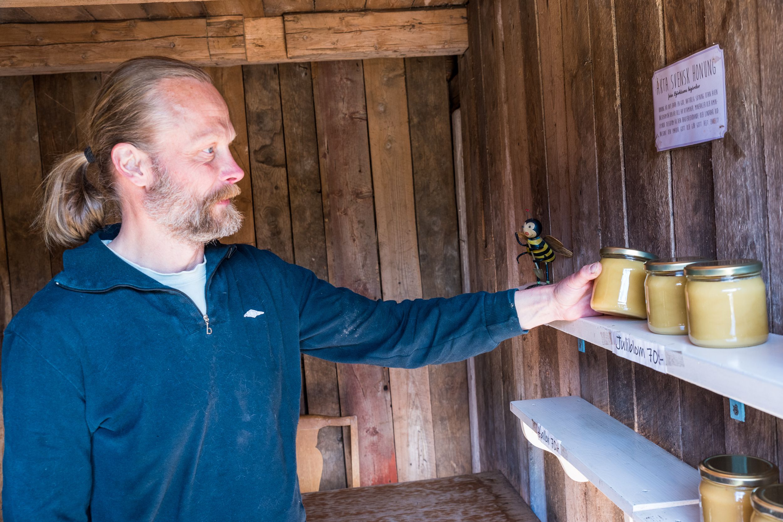En man håller i en burk honung. På väggen sitter en hylla med fler burkar honung på.