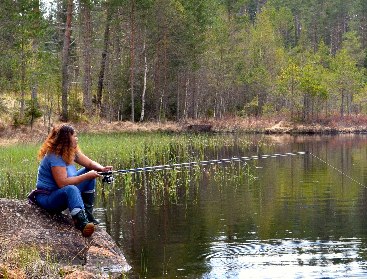 En kvinna sitter på en klippa vid sjön och fiskar. I bakgrunden skymtar man en skog.