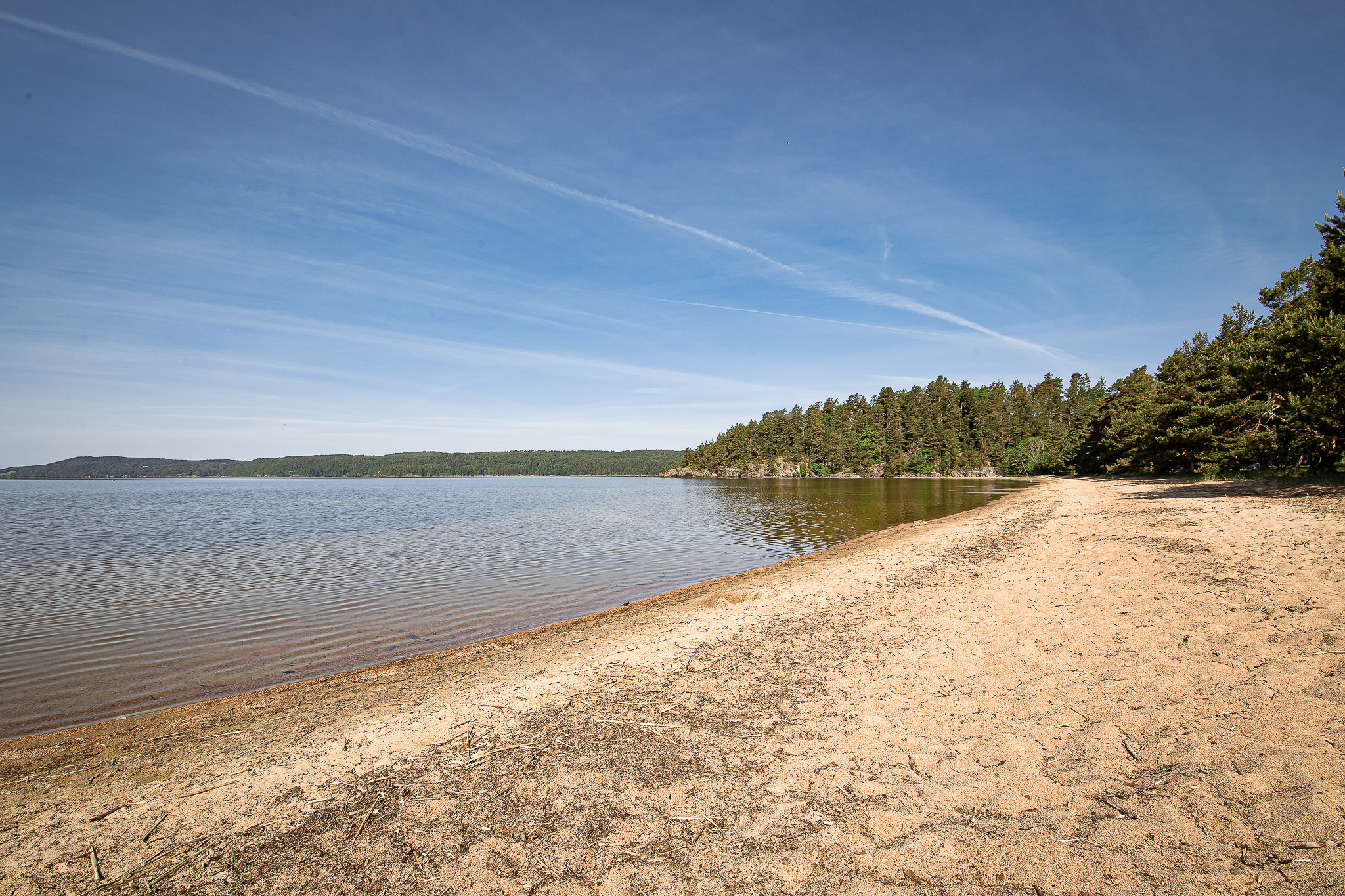 En bild på en lång och bred sandstrand som breder ut sig intill en sjö med klart vatten. Himlen är bra och i bakgrunden skymtas skog.