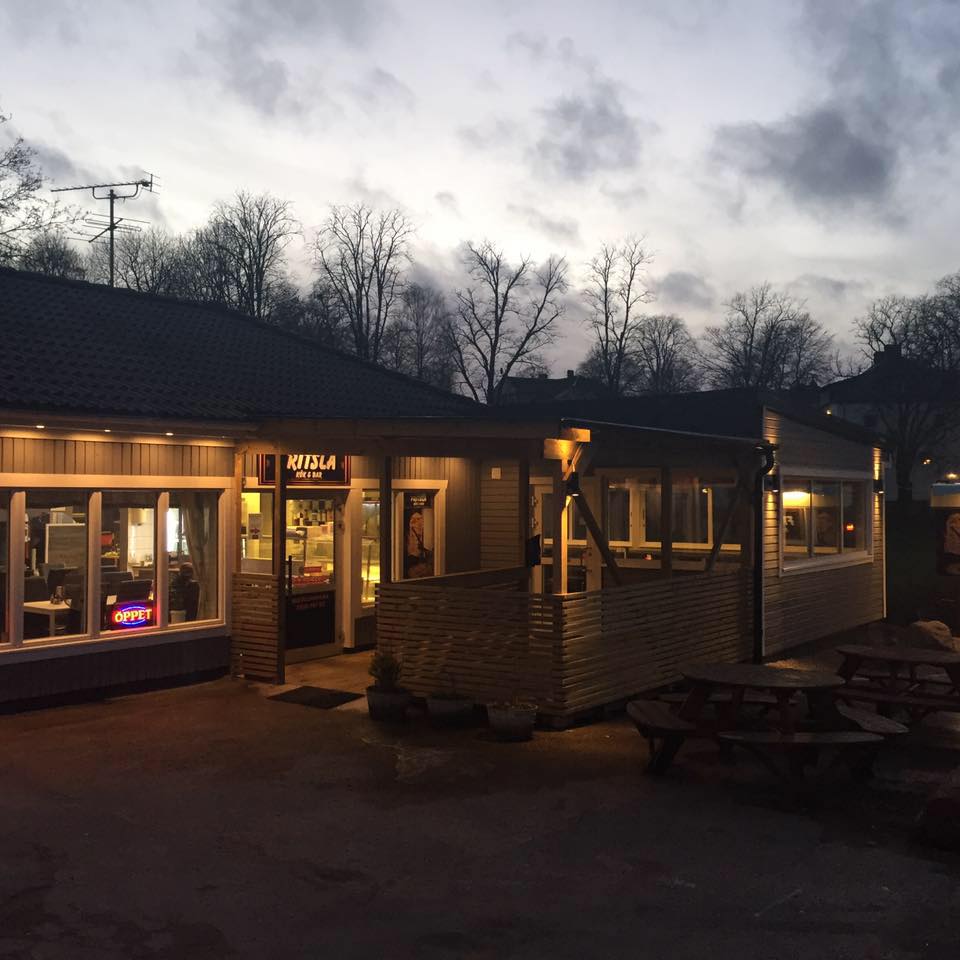 En bild på en resturang med ett trädäck för uteservering på framsidan. Här lyser många lampor, ljusslingor och neonskyltar i skymningen