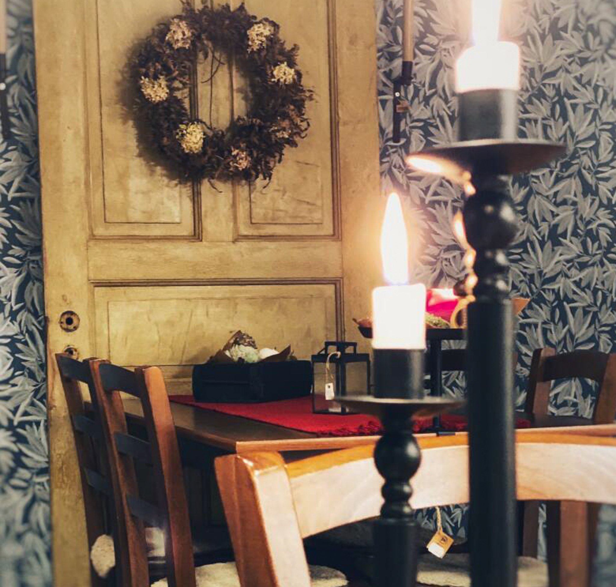 En närbild på två tända stearinljus som står i svarta ljusstakar. I bakgrunden kan man se ett fikabord med stolar. På väggen hänger en krans.