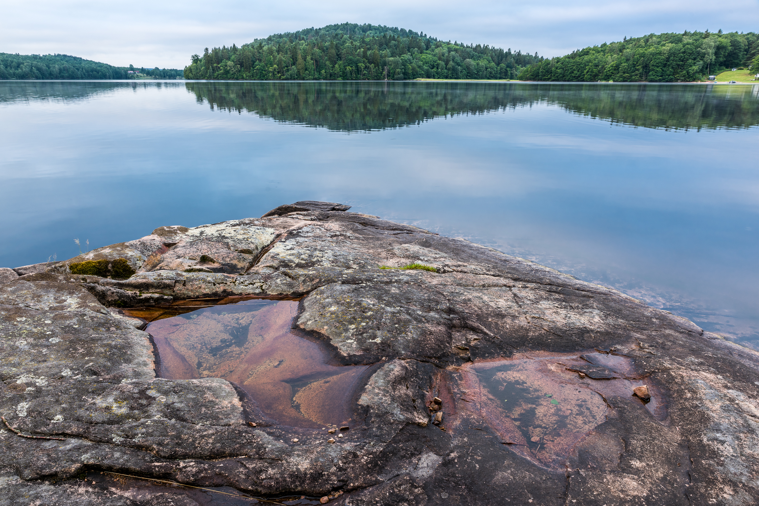En bild på en stenklippa som ligger omgivet av vatten från en stor sjö. I bakgrunden ser man en stor grön skog.
