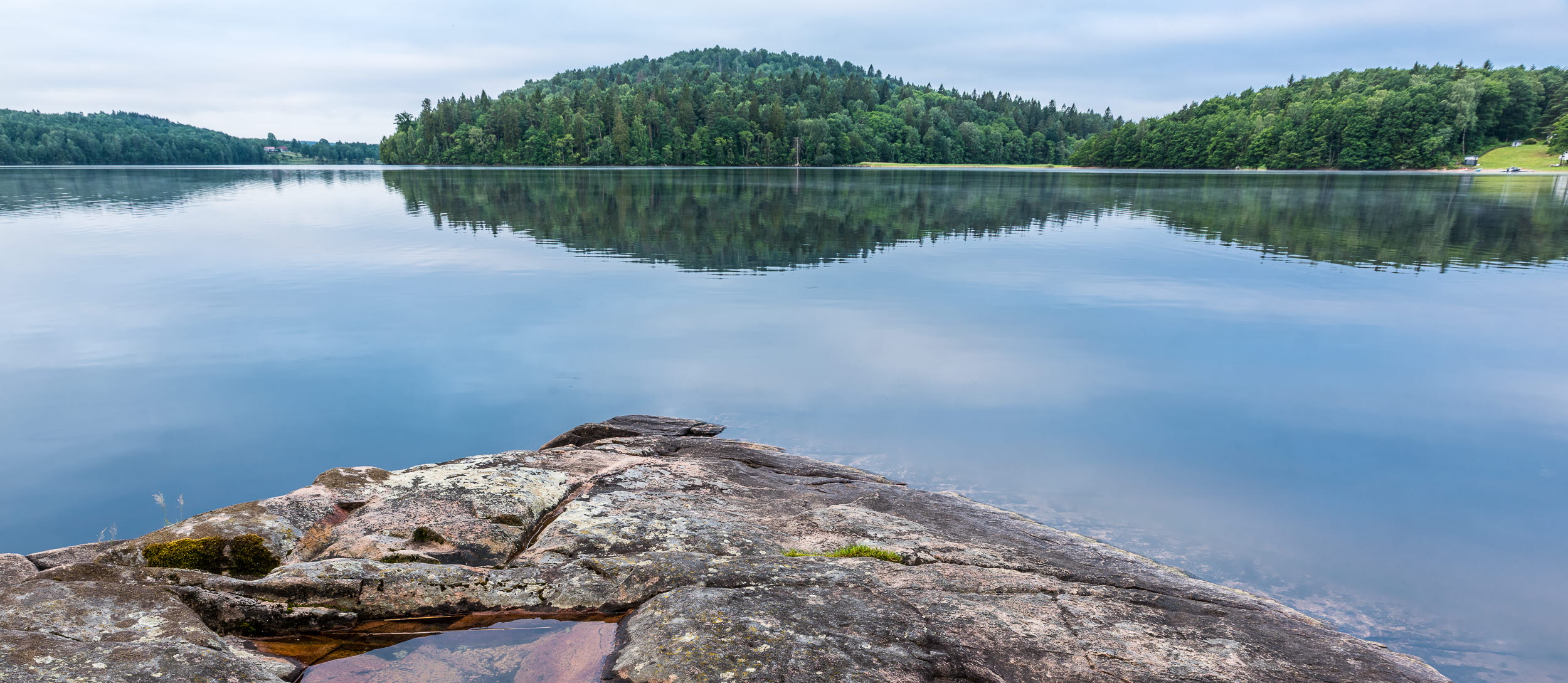 En bild på en stenklippa som ligger omgivet av vatten från en stor sjö. I bakgrunden ser man en stor grön skog.