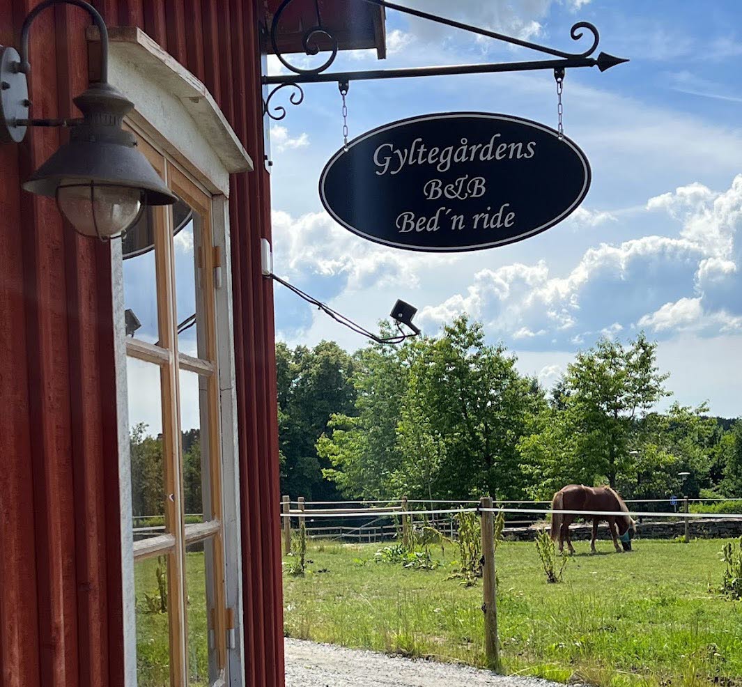 En närbild på en svart plåtskylt som hänger på en rödmålad husvägg. På skylten står det "Gyltegårdens B&B Bed' n ride"