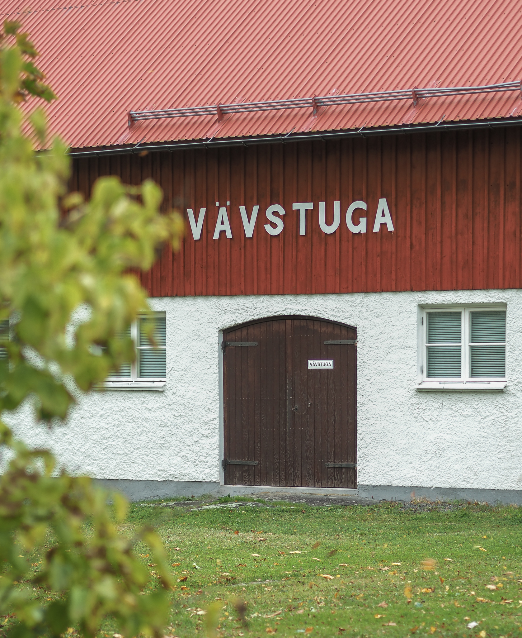 En bild på en röd ladugård med en brun port. På väggen står det "Vävstuga".