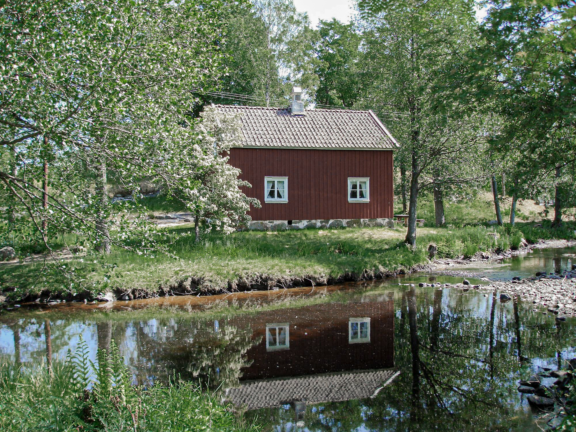 En bild på ett rödmålat hus som står i ett grönt landskap bredvid en spegelblank å.