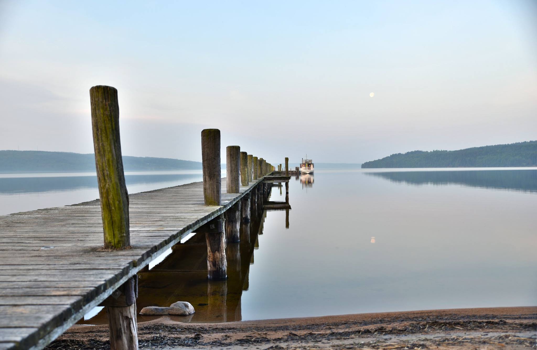En bild på en ekbrygga som leder ut till vattnet. Vid bryggans slut ligger en båt fötöjd.