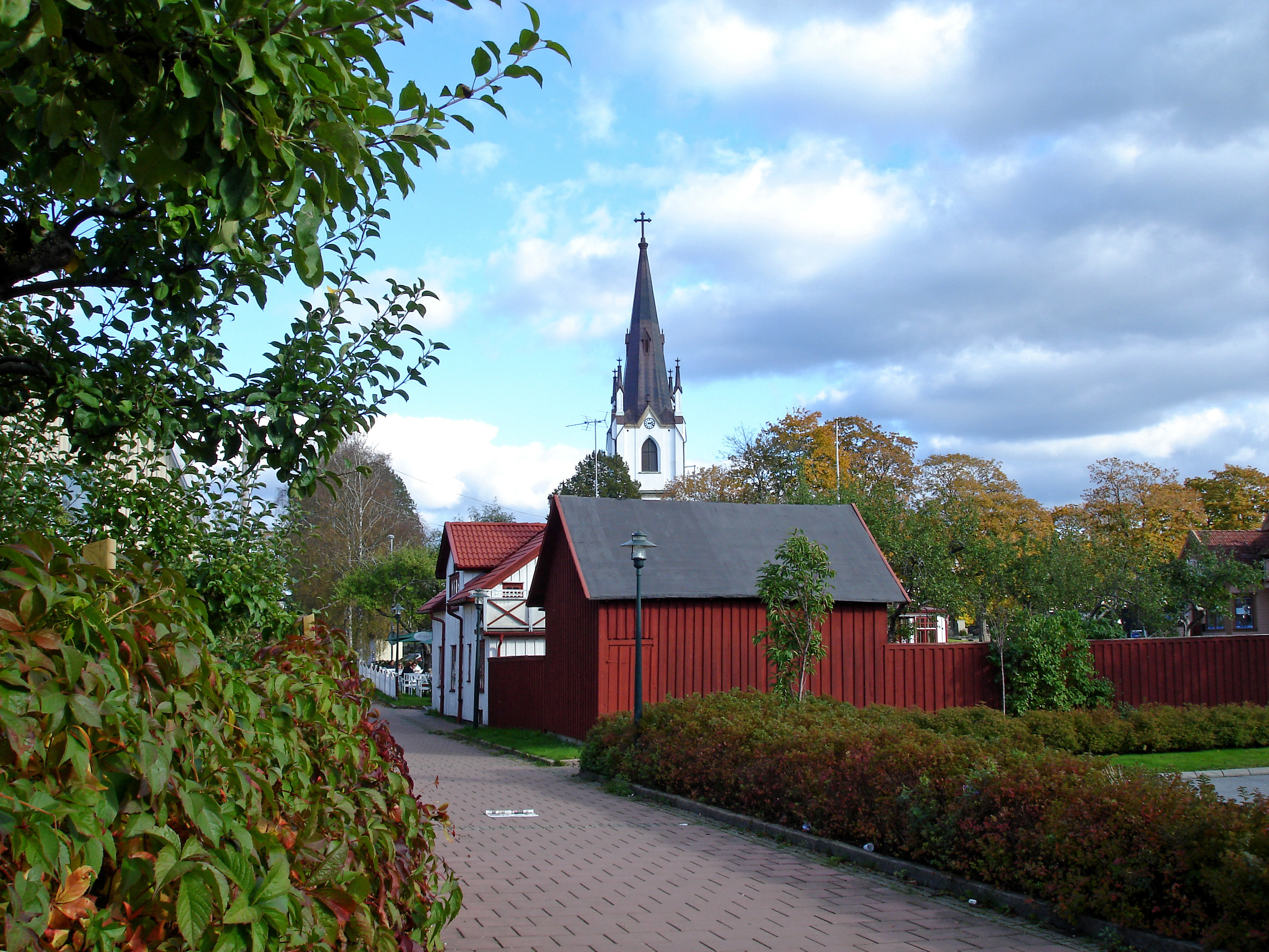 En bild på ett gångstråk i en mindre stad. Buskage och häckar ramar in en gågata och i bakgrunden ser man en kyrka och äldre trävillor.