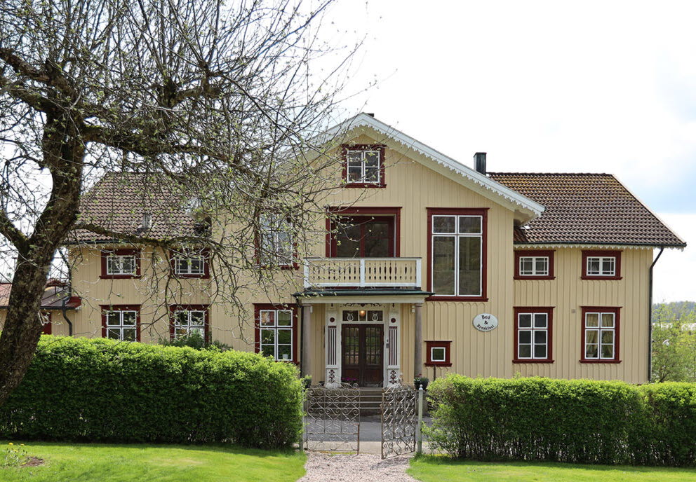 En bild på ett vackert gult trähus med röda fönsterfoder. Framför huset finns en grön häck och en vacker grusgång.