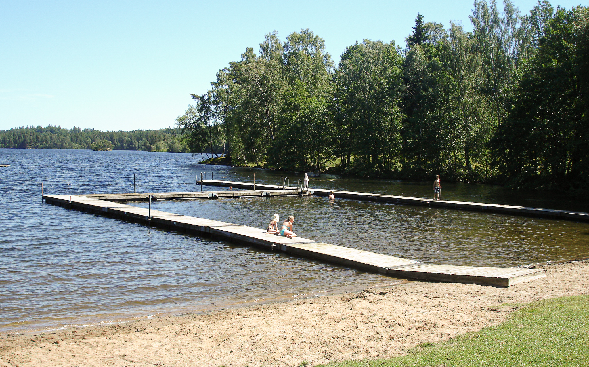 En bild på en sjö omgiven av skog. I vattnet ligger en träbrygga och några människor står på den. Intill sjön är en smal sandstrand som övergår i grönt gräs.