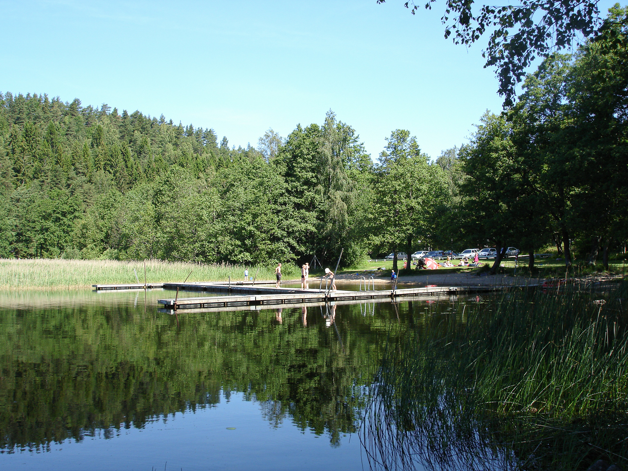 En bild på en spegelblank sjö där en lummig och grön skog speglas i den blå vattenytan. I vattnet ligger en träbrygga och det står ett par människor på den. Himlen är blå och det är soligt.
