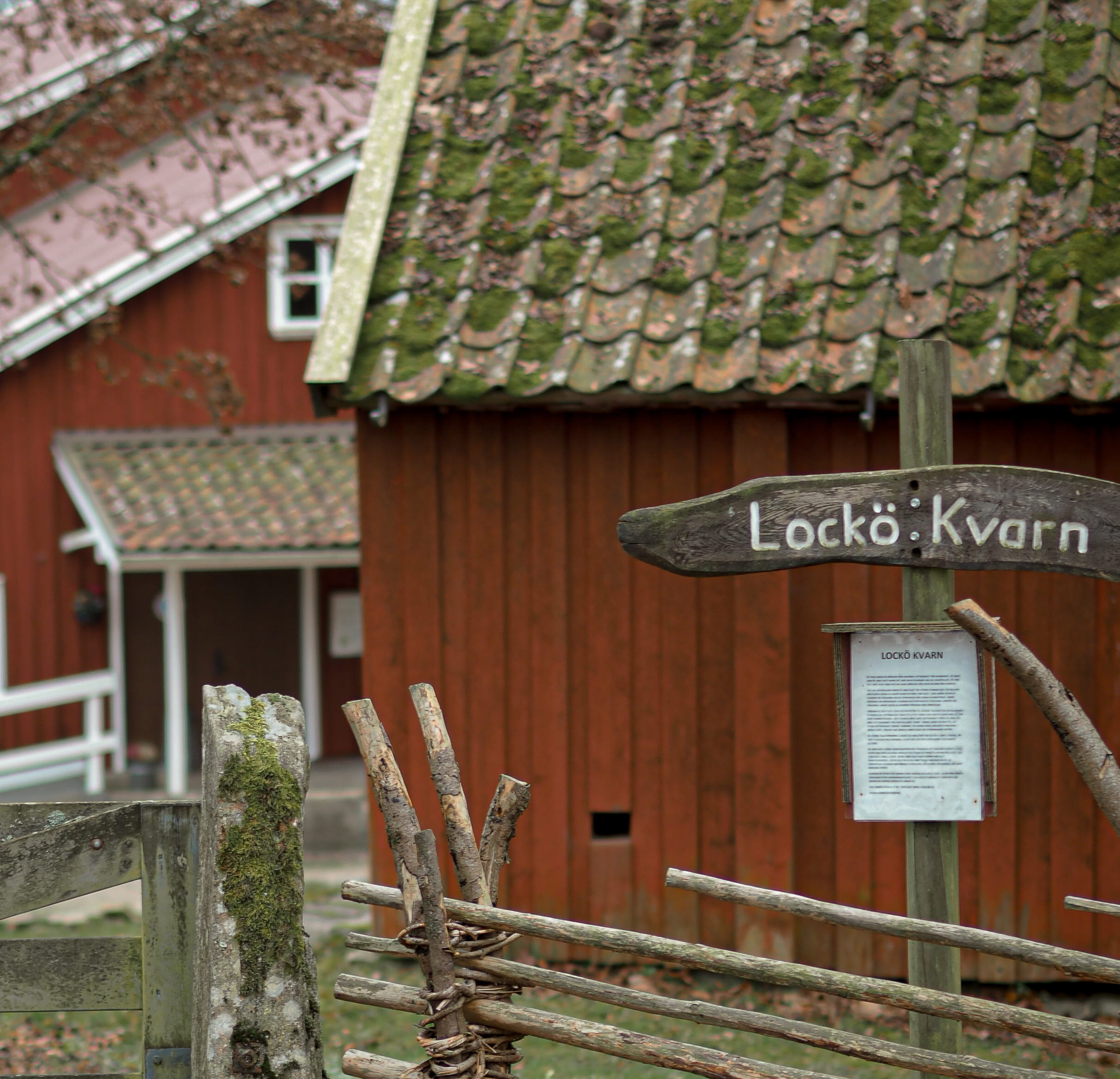 En bild på ett staket med en skylt där det står "Lockö kvarn". I bakgrunden ligger rödmålade hus.