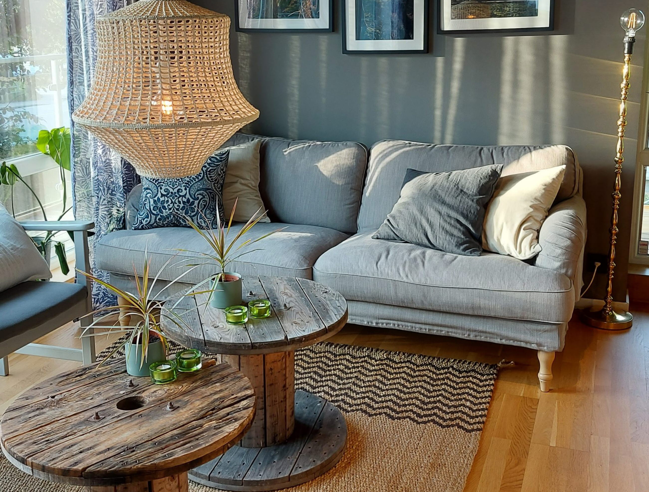 En bild på en soffa med prydnadskuddar som står mot en mörk vägg med tavlor på. Framför soffan står två runda bord. Solen lyser in genom ett fönster. Från taket hänger en stor taklampa.