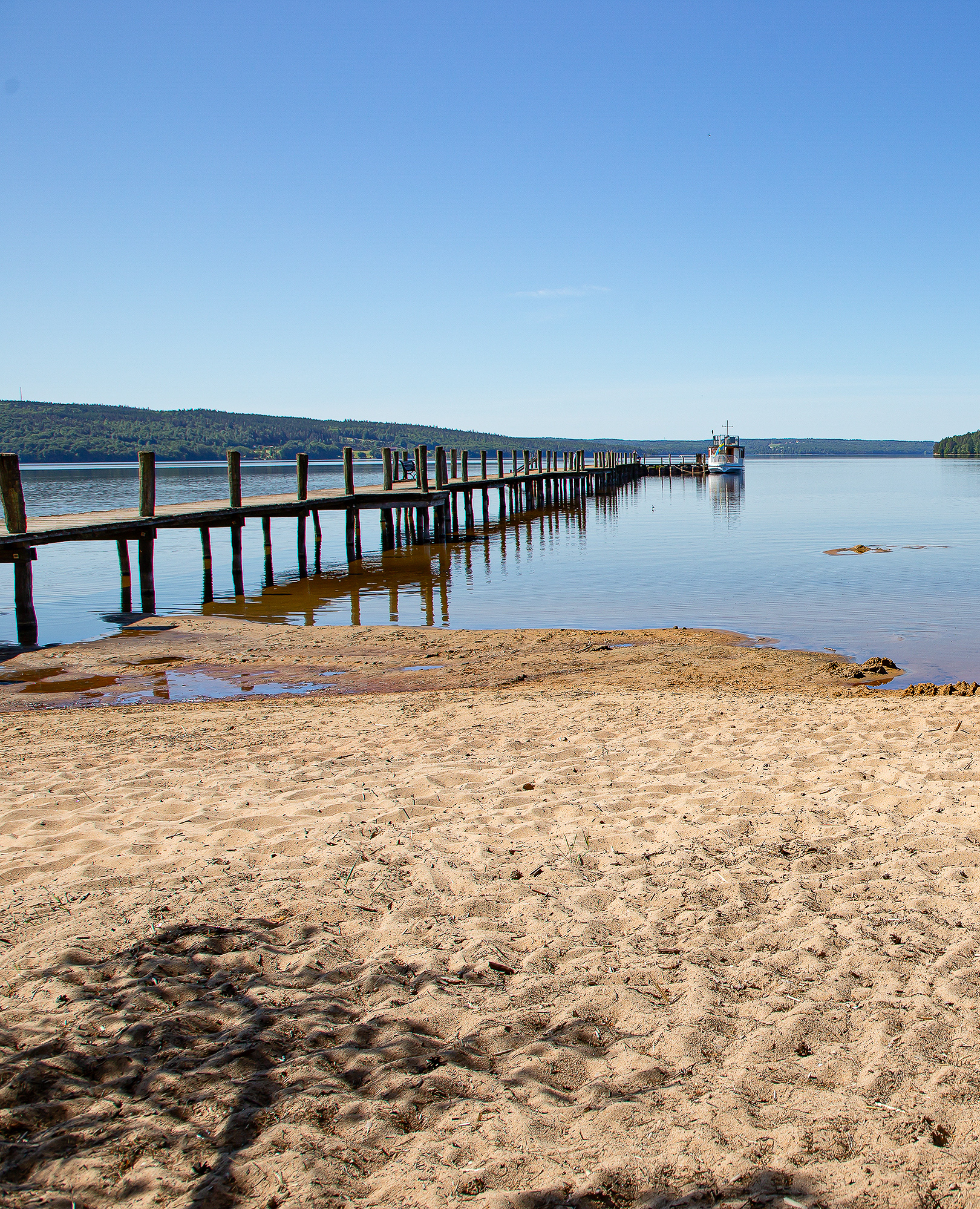 En bild på en sandstrand intill en stor blå sjö- Himlen är klarblå och molnfri. En stor, rejäl brygga går rakt ut i vattnet. Längst ut på bryggan ligger en båt.