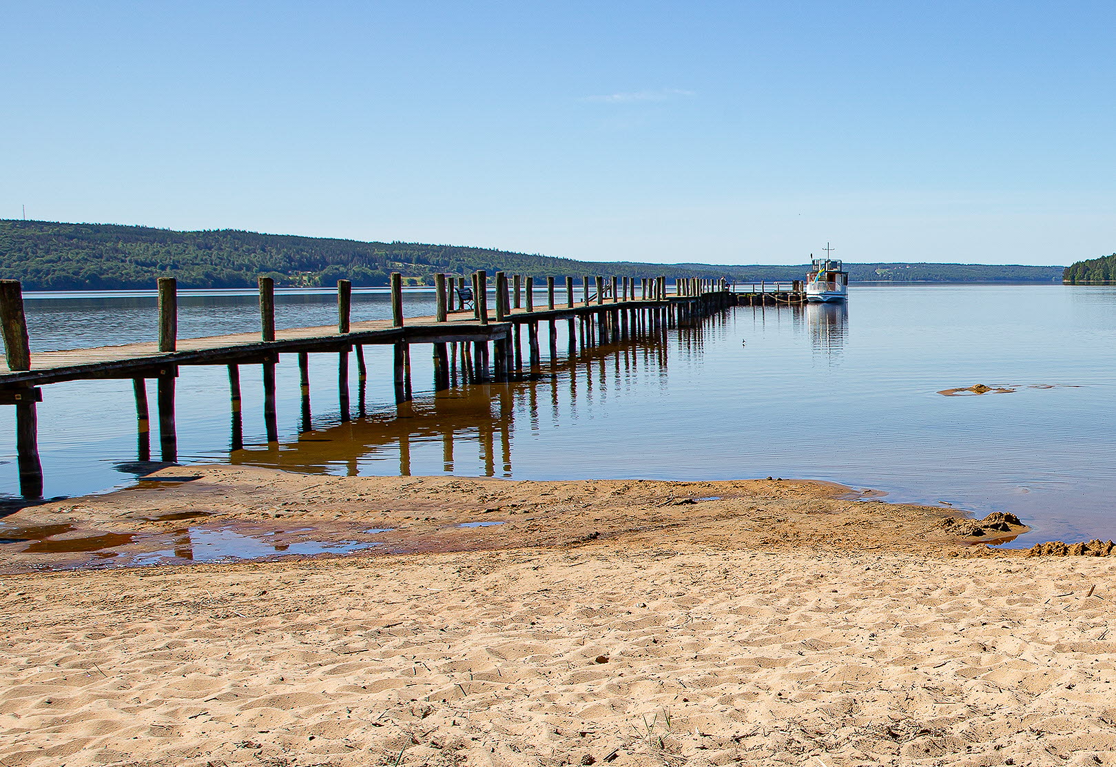 En bild på en sandstrand intill en stor blå sjö- Himlen är klarblå och molnfri. En stor, rejäl brygga går rakt ut i vattnet. Längst ut på bryggan ligger en båt.