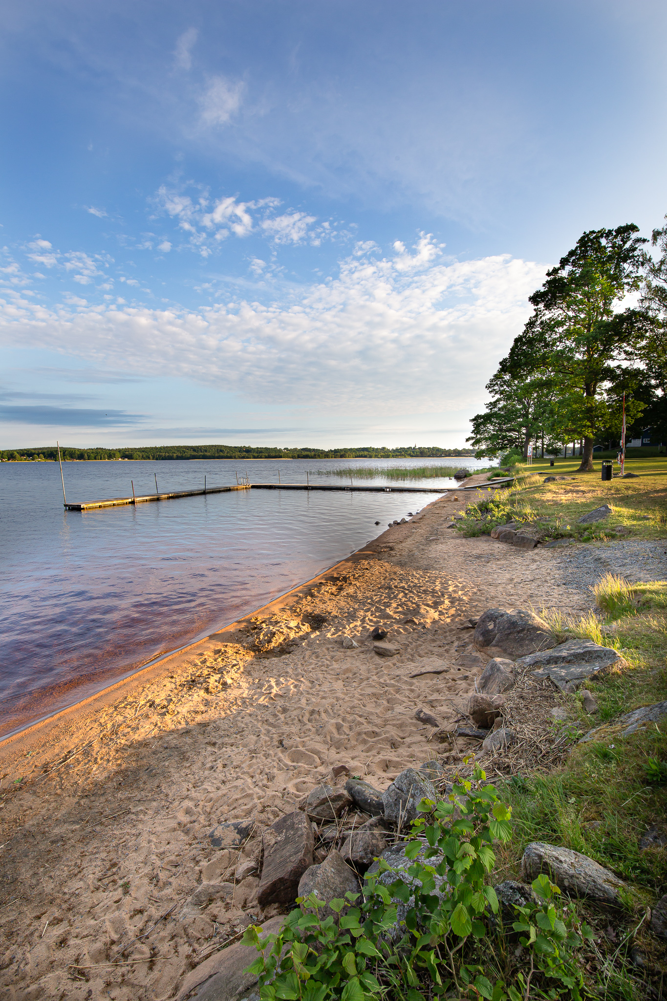 En bild på en smal sandstrand som kantas med stenbumlingar och lite växtlighet. Intill stranden ligger en sjö med en brygga i.