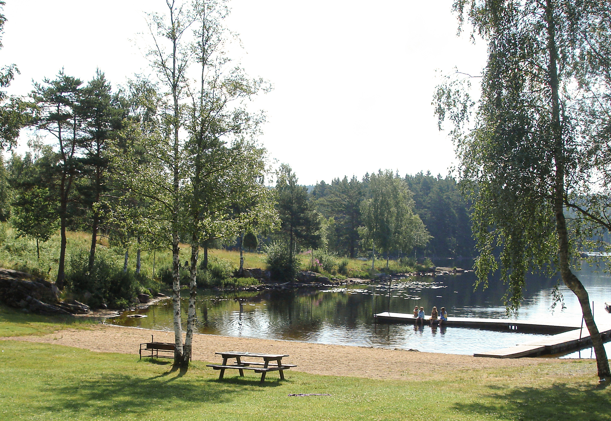 En bild på en liten sjö omgiven av skog. En sandstrand och en gräsplätt ligger intil sjön. Här står en bänk på gräset och i vattnet ligger en brygga.