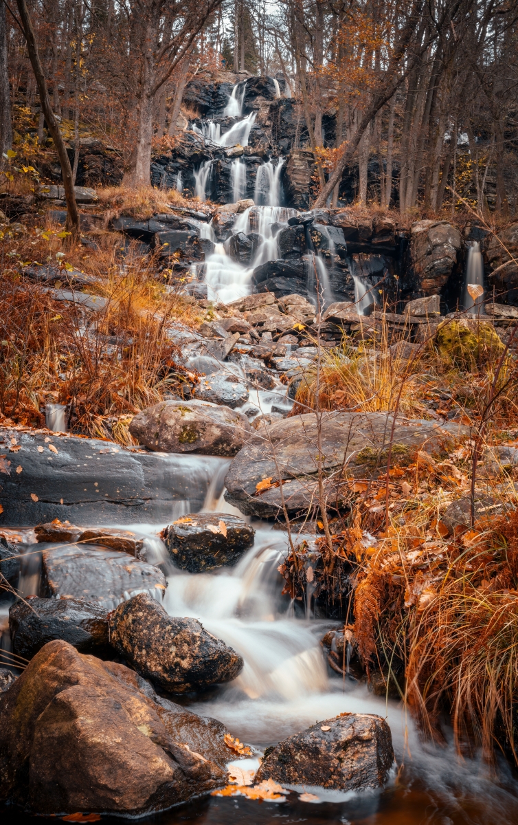 En bild på ett vattenfall där vatten rinner ner för stenklippor omgivet av ett höstligt, orangefärgat, landskap.