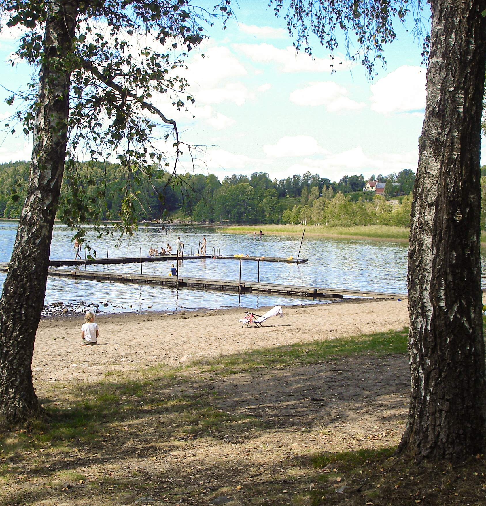 Mellan två björkar kan vi skymta en sandstrand och en liten sjö med en brygga i. På bilden förekommer flera badsugna människor denna härliga sommardag.