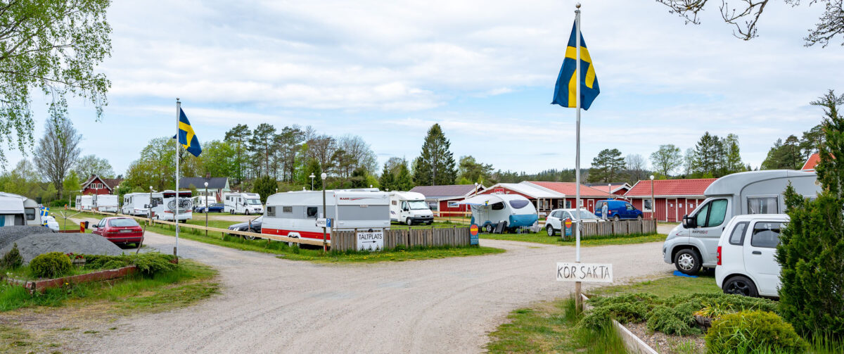 En bild tagen från infarten till en campingplats där mängder av husvagnar och husbilar står uppradade på en gräsmatta. Små rödmålade stugor och en del Sverigeflaggor syns också.