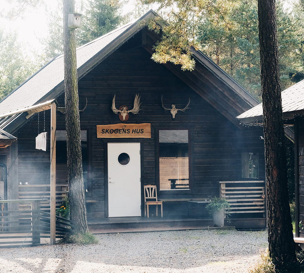 En bild på tre svartmålade små stugor som stor omringade av frodig skog.