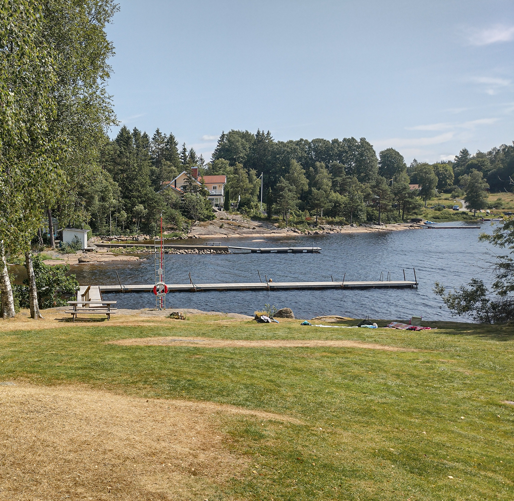 Intill en stor gräsmatta ligger en sjö med härligt blått vatten. I sjön ligger en brygga. I bakgrunden skymtas skog och hus.