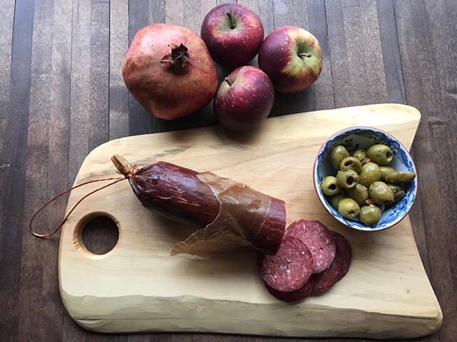 En bild på ett bord med en skärbräda där det ligger en rejäl korv som någon skurit upp några skivor av. På skärbrädan står också en liten porslinsskål med oliver. Jämte skärbrädan ligger röda äpplen och ett granatäpple.