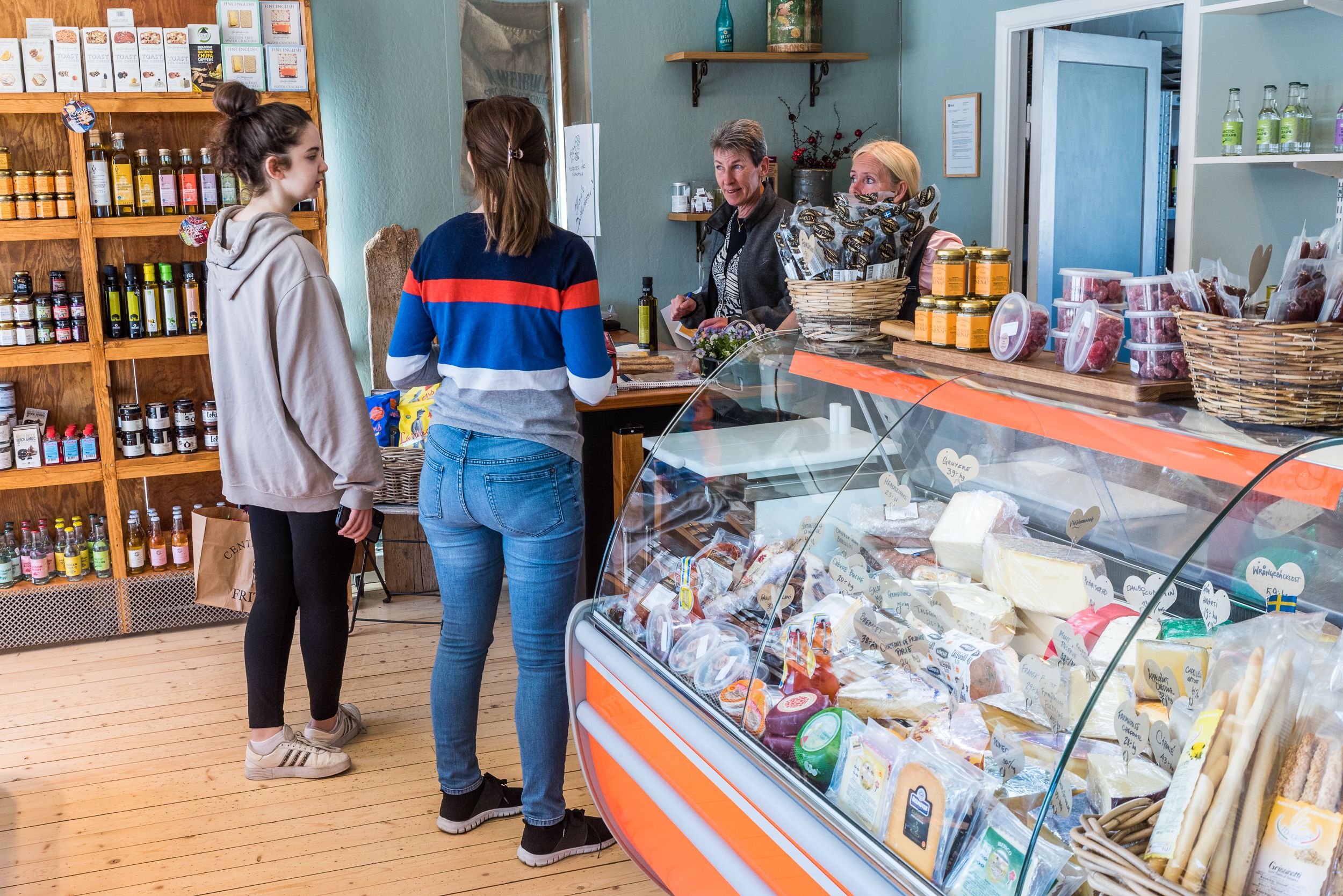 Två kvinnor står och handlar över disk i en delikatessbutik. En tredje kvinna står bakom disken och tar betalt.