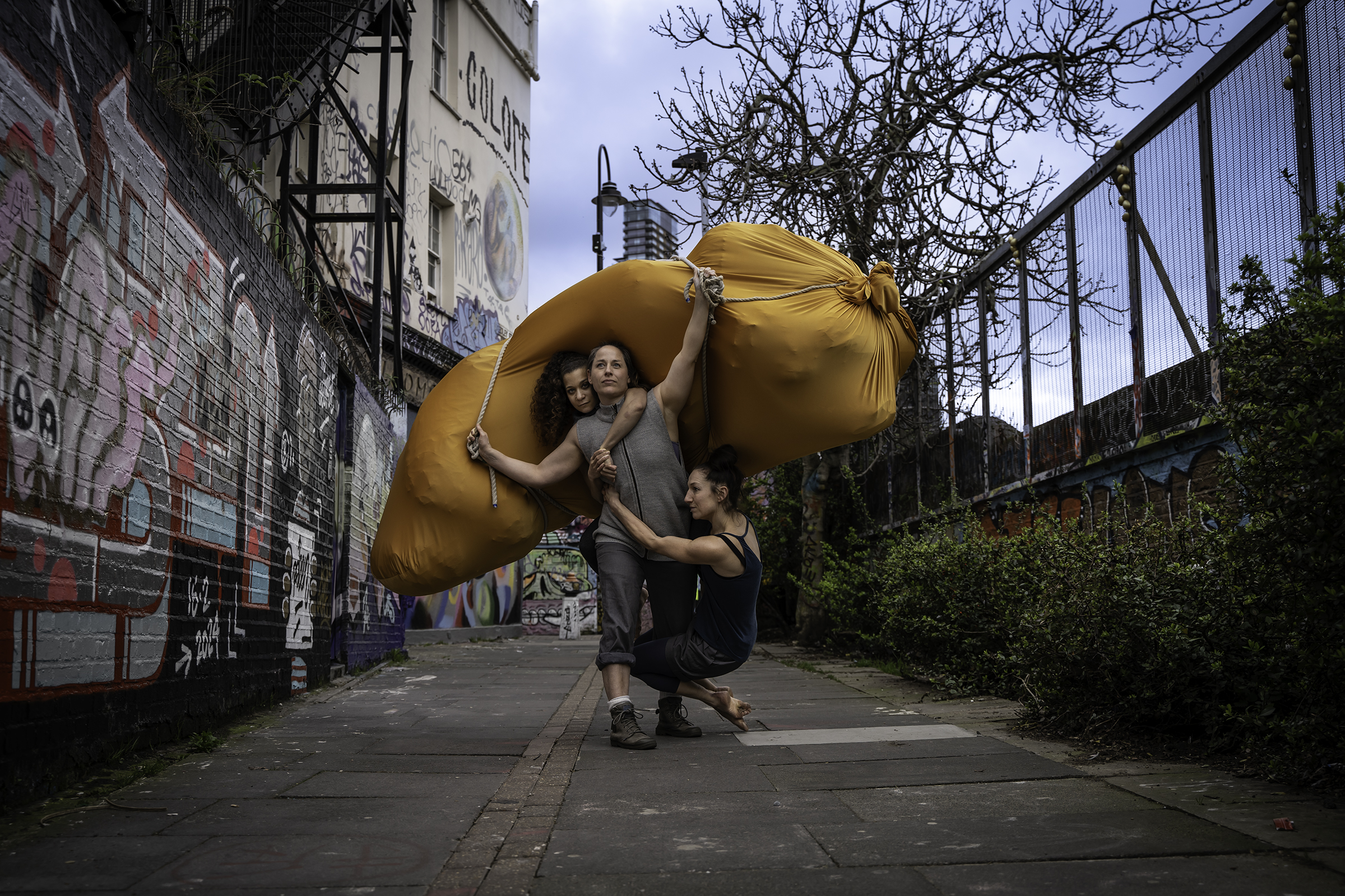 Tre kvinnor i grå färgade kläder utför ett akrobatiskt balans trick i en gränd. 