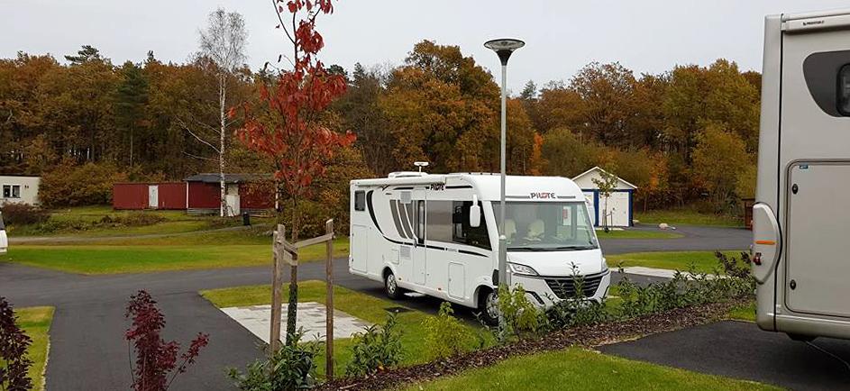 En vit husbil står parkerad på en ställplats och i omgivningen syns träd i höstfärger.