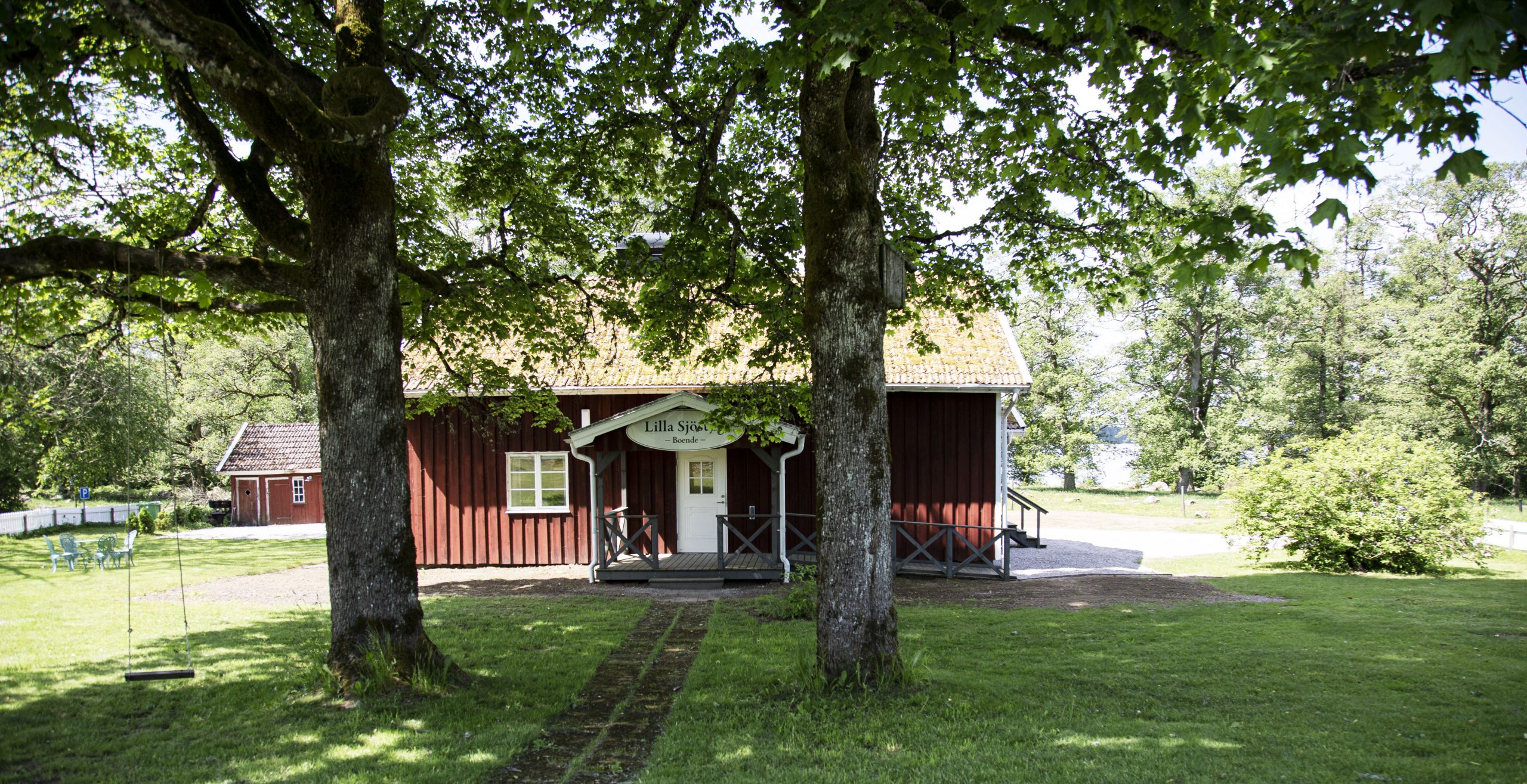 Klassisk svensk stuga i sommaridyll. Röd stuga med vita knutar bagom två lövträd där det hänger en gunga i, grön gräsmatta.