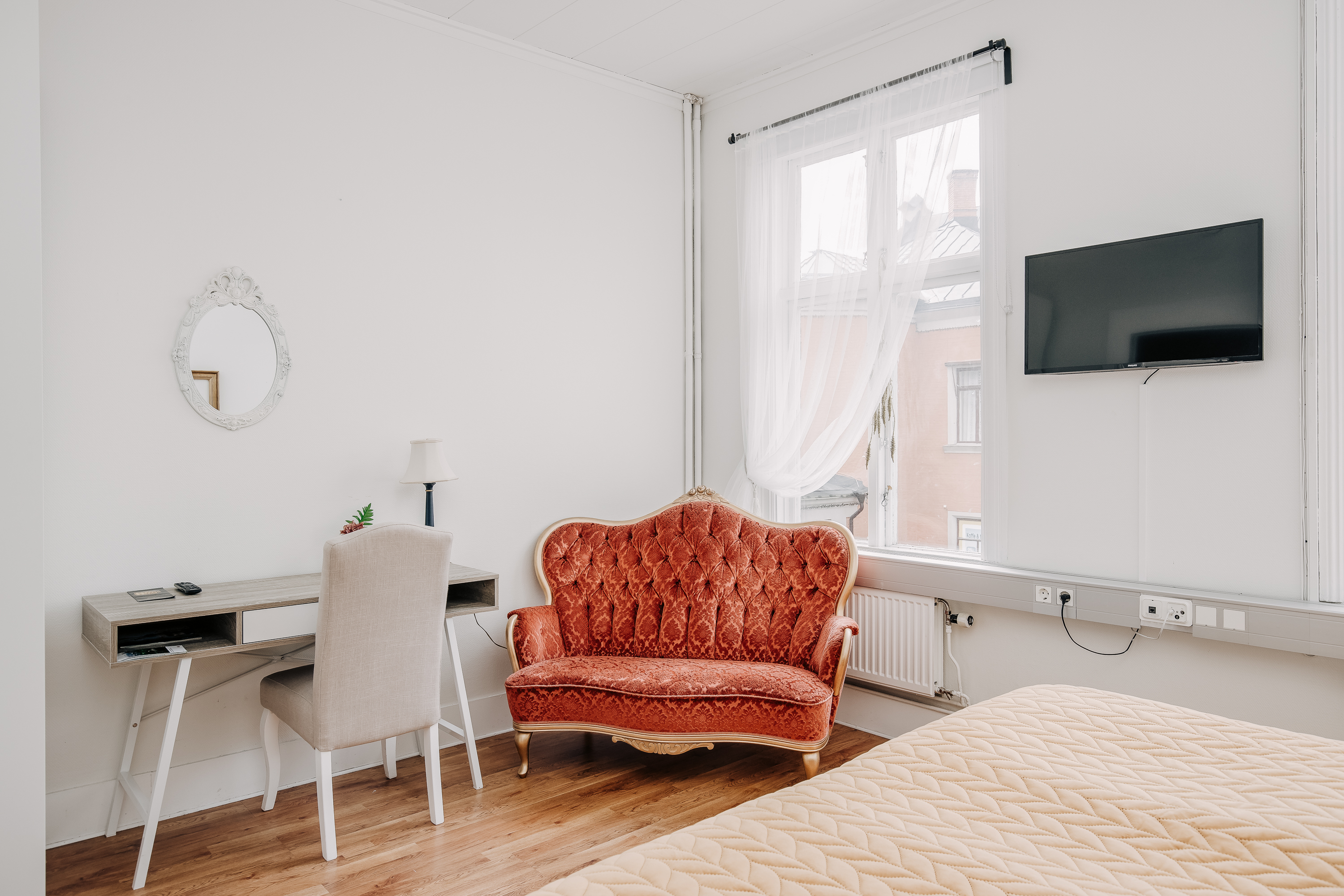 A bright hotel room. In the room there is a small desk, a red rococo couch and a tv. 