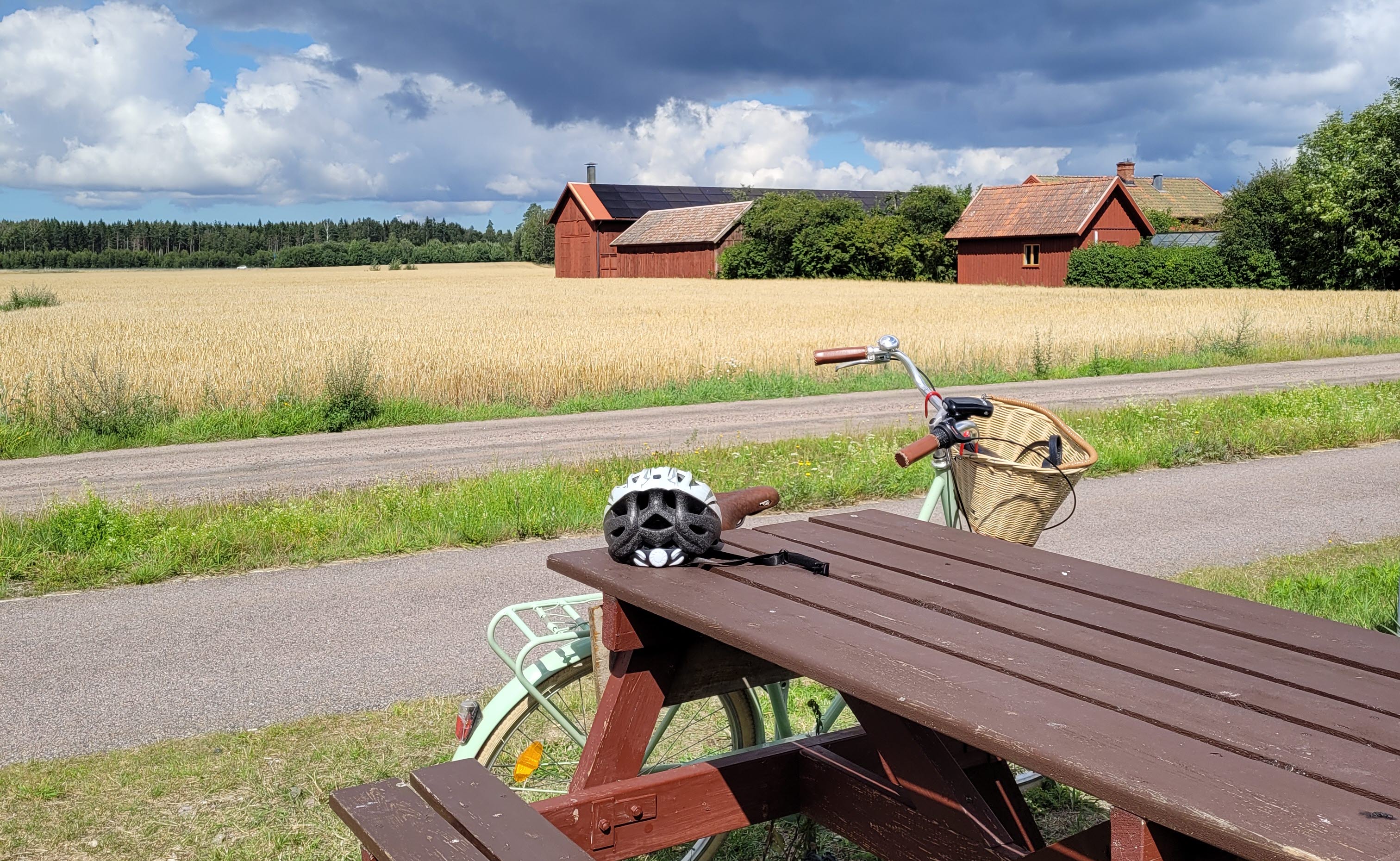 En cykel står lutad mot ett picknickbord. I bakgrunden syns en gård med röda ladugårdar.