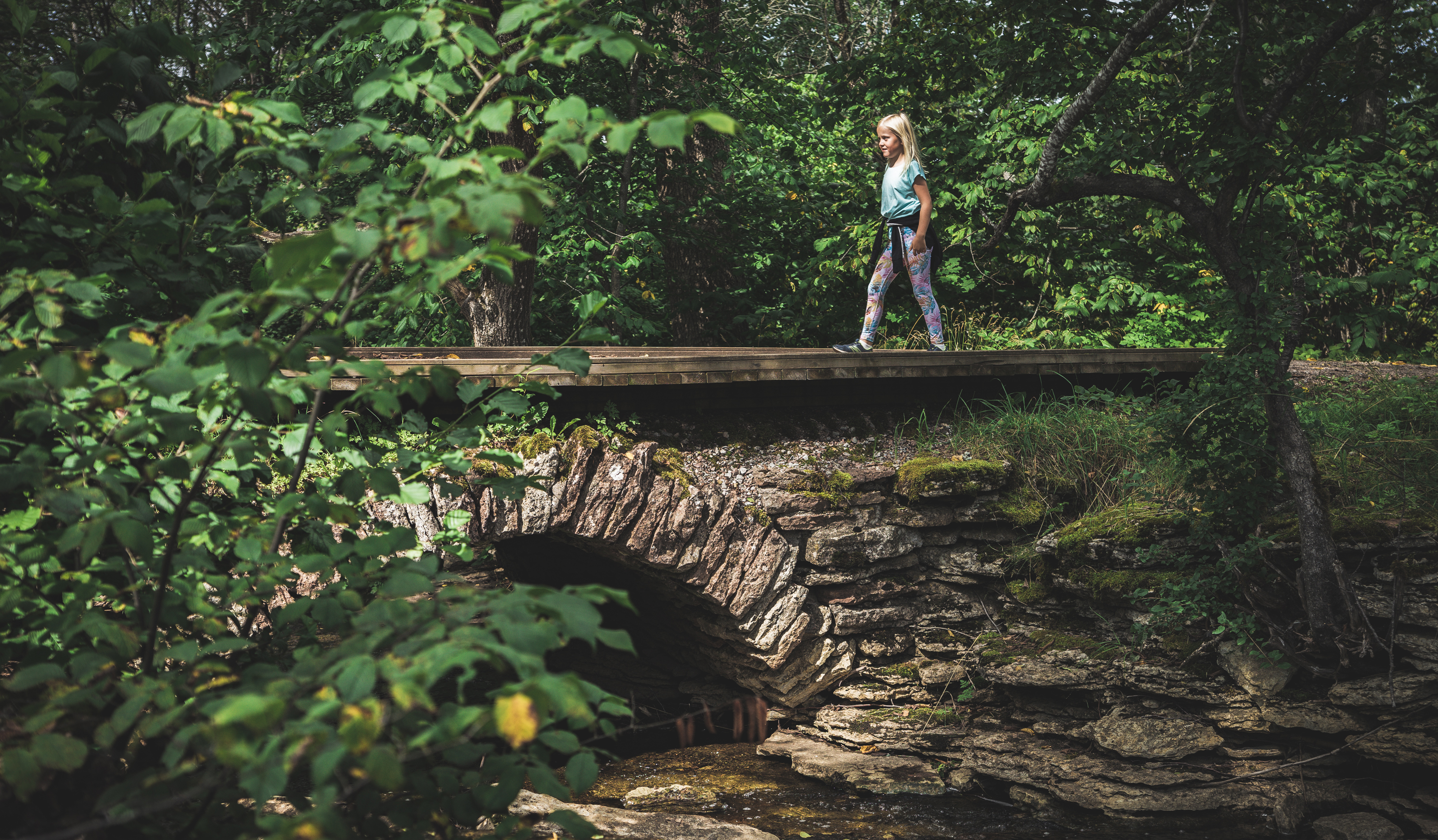 Ett barn går över en välvd stenbro. Omgivningarna är gröna och lummiga. 