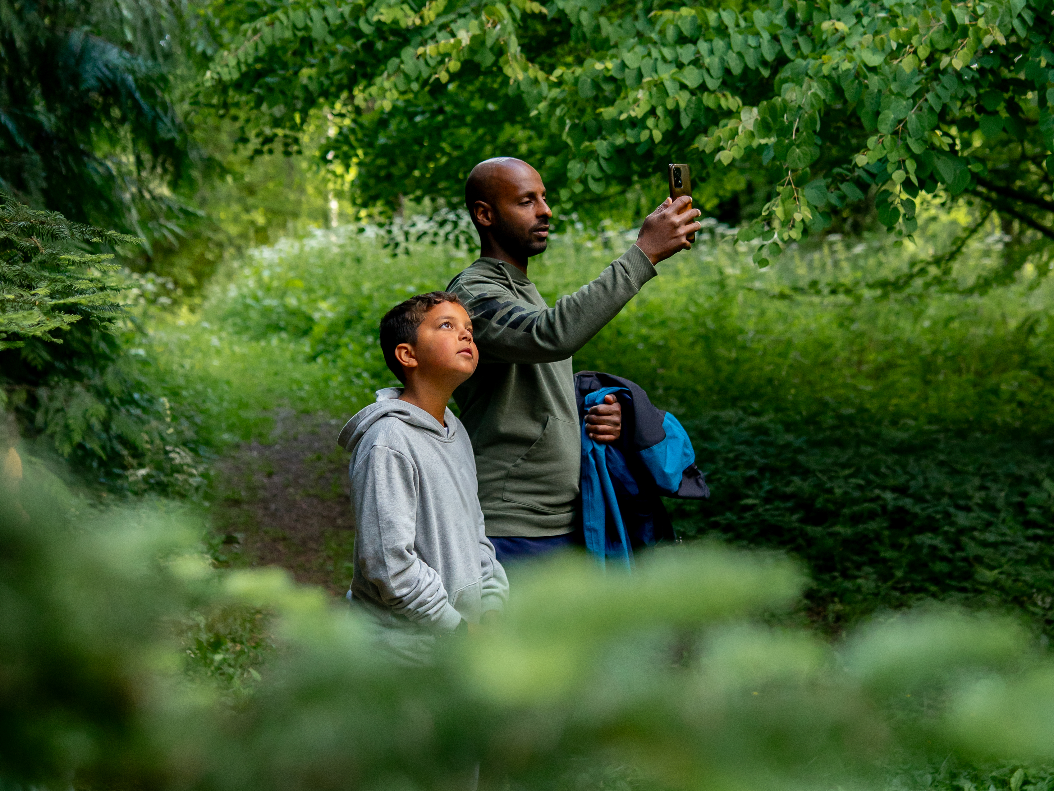En pappa och ett barn tittar på gröna träd. Pappan fotar med sin telefon.