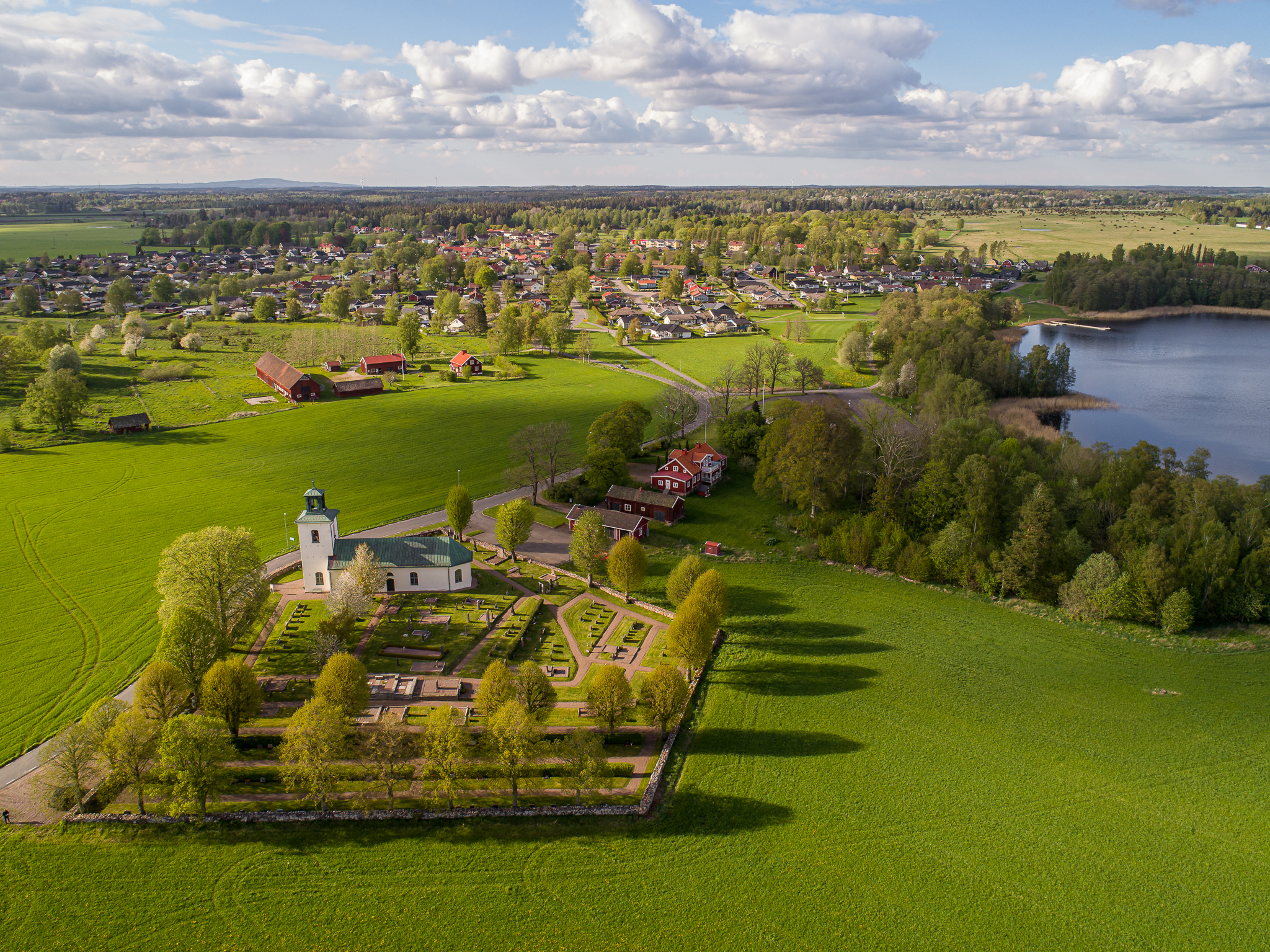 Drönarbild över grönskande fält, sjö och vit kyrka. 