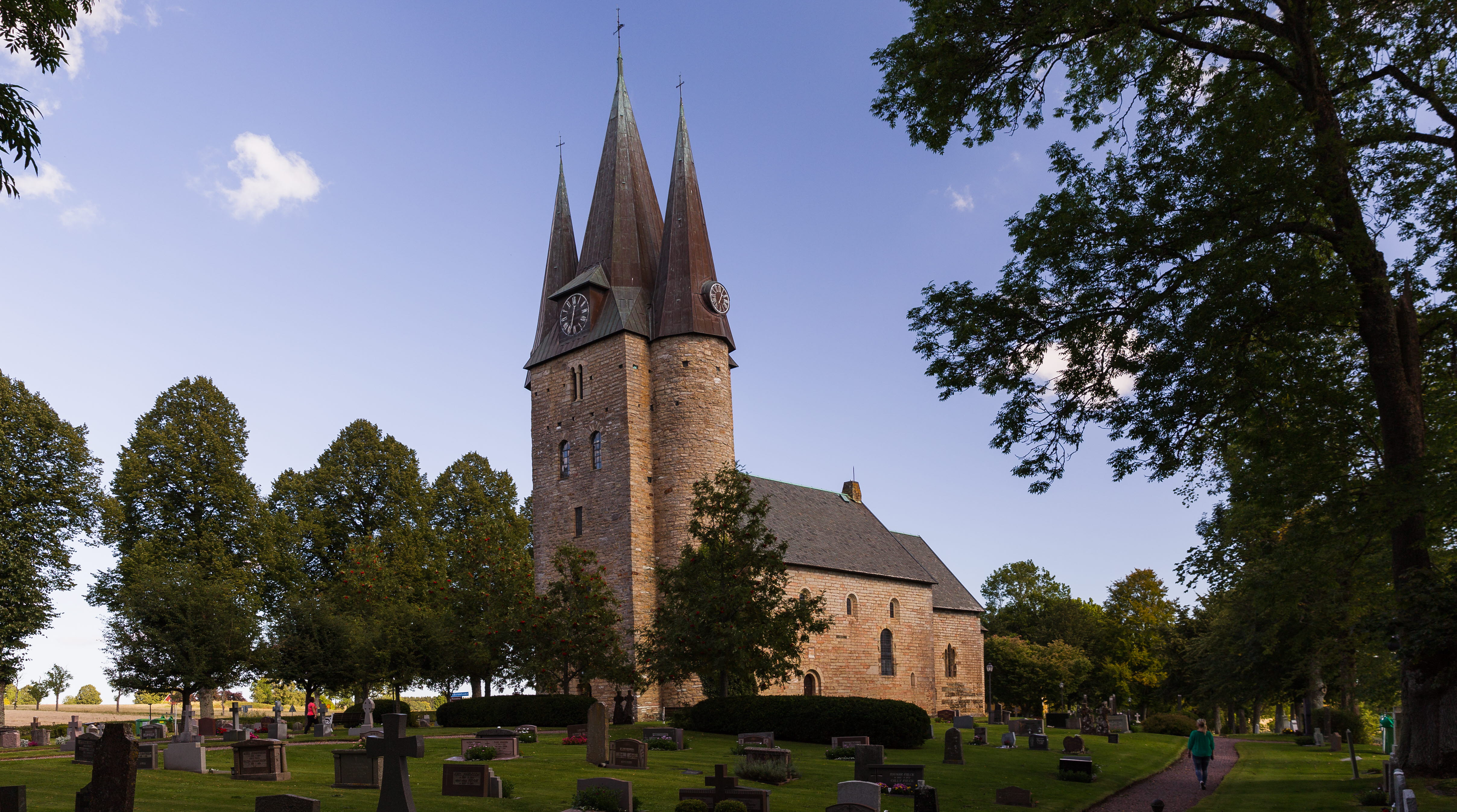 Foto på den medeltida kyrkan Husaby kyrka. I förgrunden finns gröna träd. 